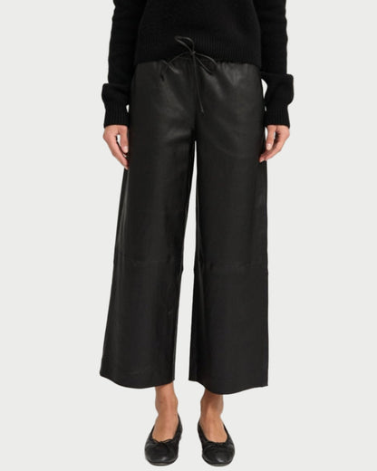 A person wearing SPRWMN Cropped Leather Baggy Pants, a black long-sleeve top, and black flat shoes stands against a plain light background.