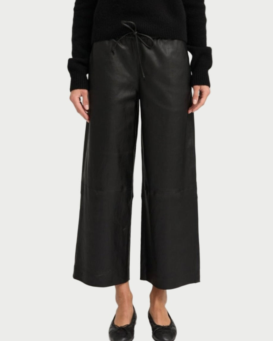A person wearing SPRWMN Cropped Leather Baggy Pants, paired with a black long-sleeve sweater and black flats, stands against a plain light background. Only the figure from the shoulders down is visible.