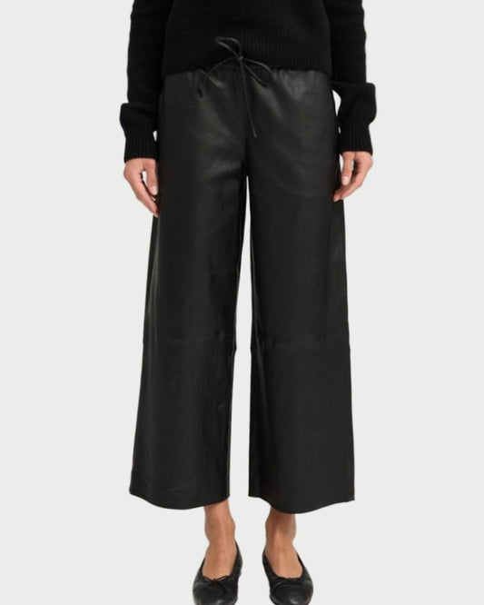 A person wearing SPRWMN Cropped Leather Baggy Pants, paired with a black long-sleeve sweater and black flats, stands against a plain light background. Only the figure from the shoulders down is visible.