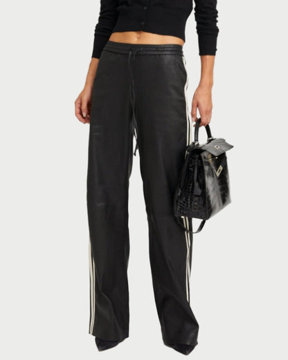 A person wears SPRWMN Baggy Athletic Pants—black with white side stripes—a black cropped cardigan, and holds a black textured handbag. Head and feet are not visible.