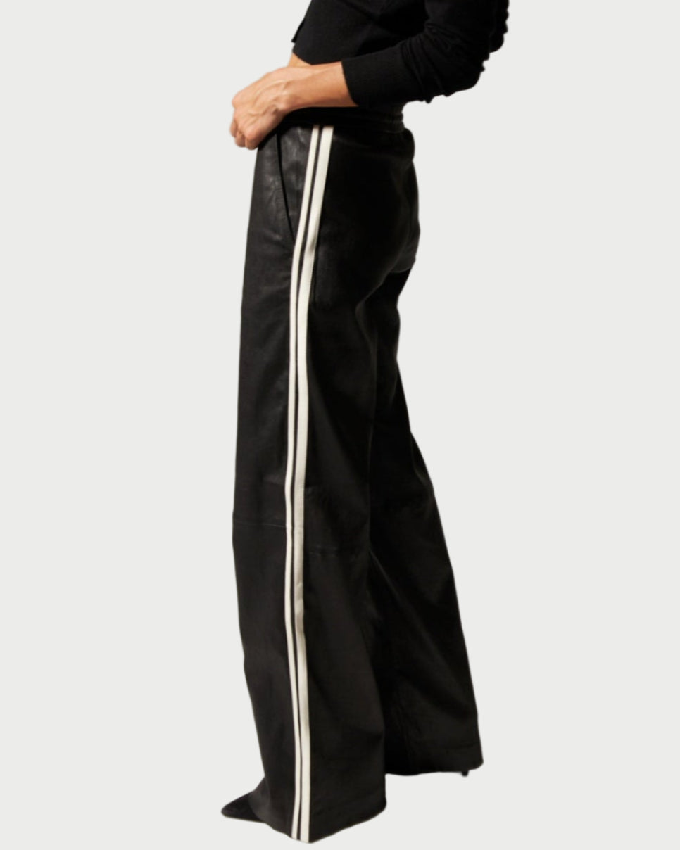 A person wears SPRWMN Baggy Athletic Pants—high-waisted, wide-leg track pants with white side stripes by SPRWMN—and a black long-sleeve top, standing against a light background.
