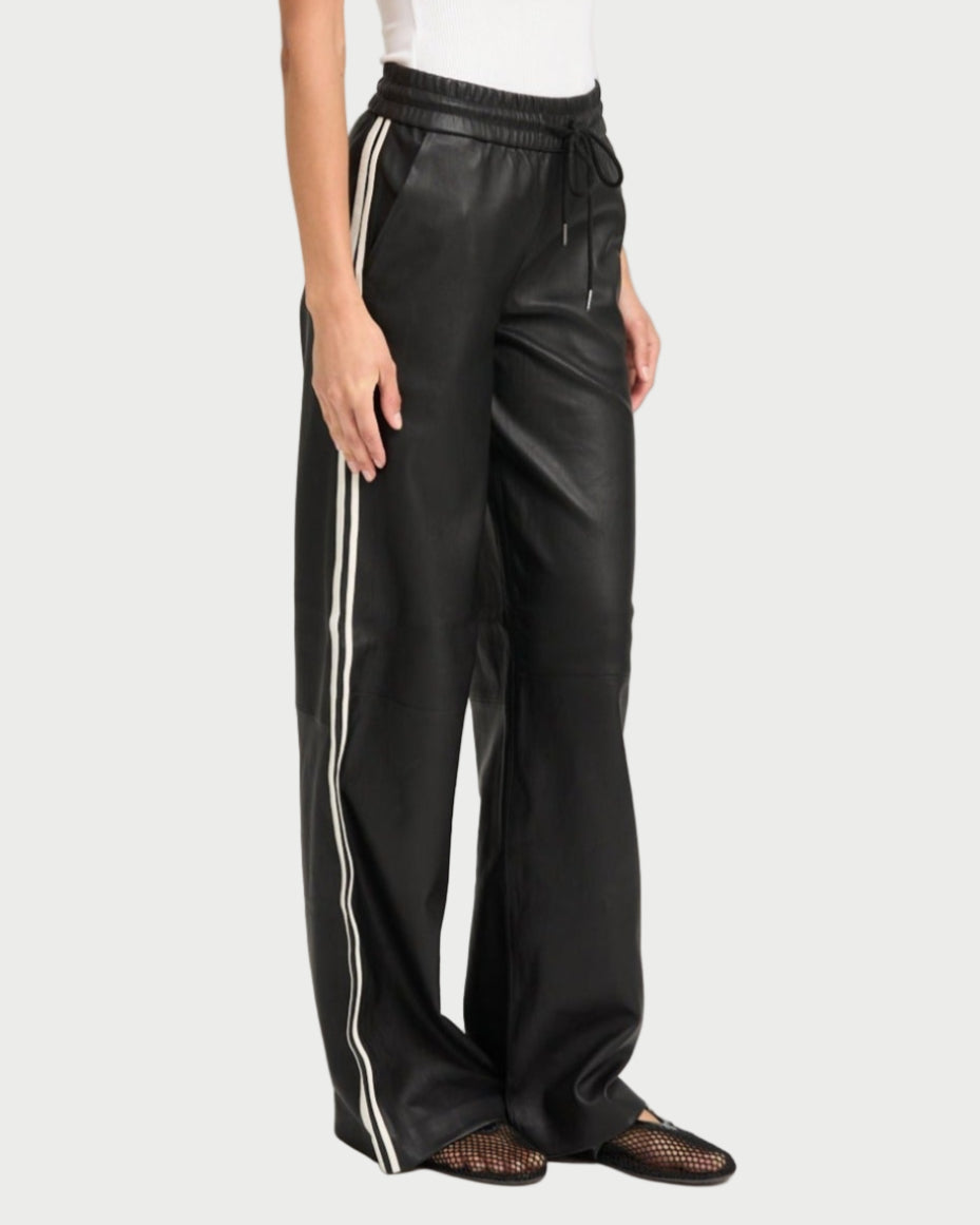 Someone wearing SPRWMN Baggy Athletic Pants by SPRWMN—black, wide-leg with white side stripes and an elastic drawstring waist—paired with a white sleeveless top and black mesh shoes stands against a plain light background.