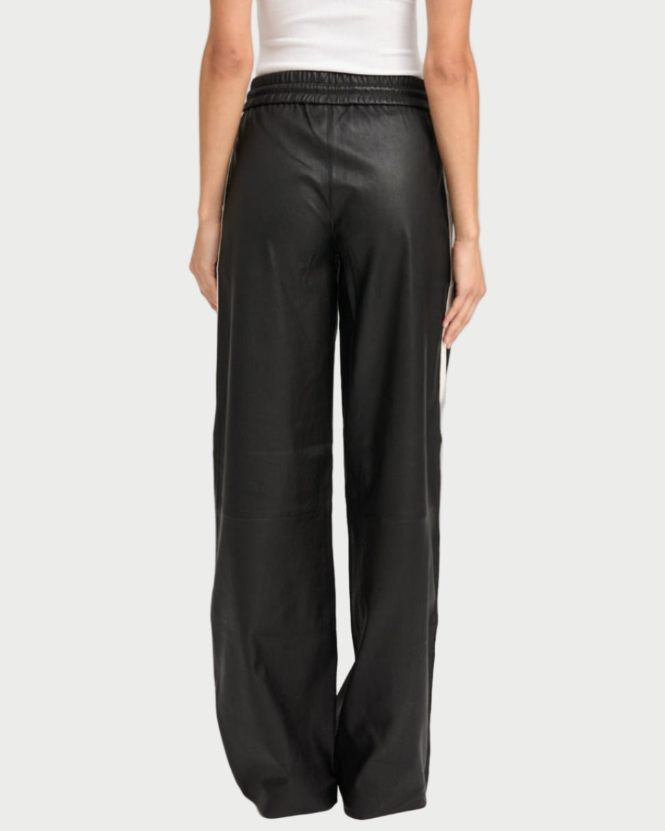 A person is shown from the back, standing against a plain light background, wearing SPRWMN Baggy Athletic Pants in black leather paired with a white sleeveless top.