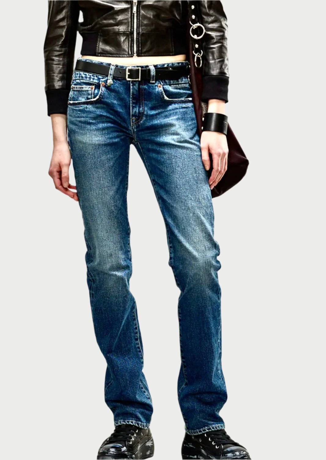 A person stands against a plain background wearing a black leather jacket, R13 Tobi Boy Jean by R13 in selvedge blue, a white belt, black shoes, and a black wristband, with one hand by their side and a bag strap on their shoulder.