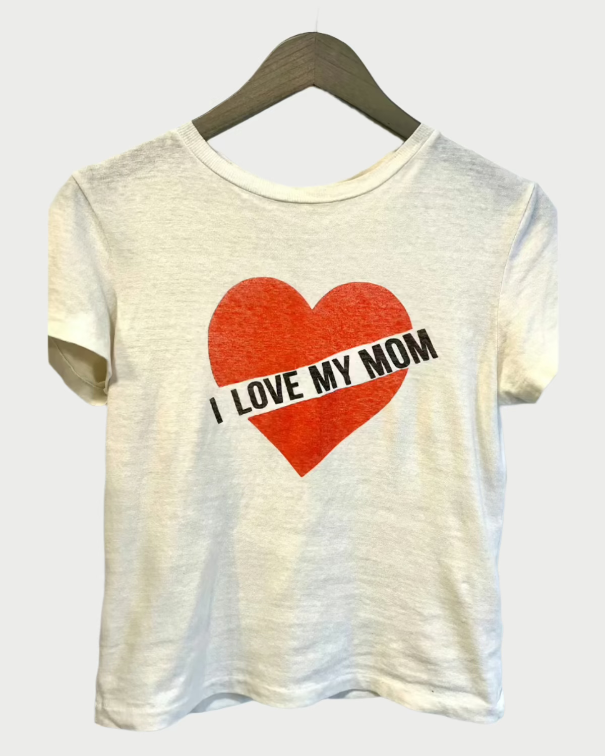 The "I Love My Mom T-Shirt" by RE/DONE showcases a bold red heart graphic on the front with "I LOVE MY MOM" text inside. Made from 100% cotton, this T-shirt is displayed on a wooden hanger against a plain backdrop, perfectly combining timeless style with heartfelt sentiment.