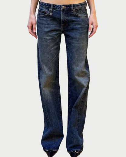 A person wearing dark blue straight-leg jeans with a faded wash, standing against a white textured wall on a wooden floor.