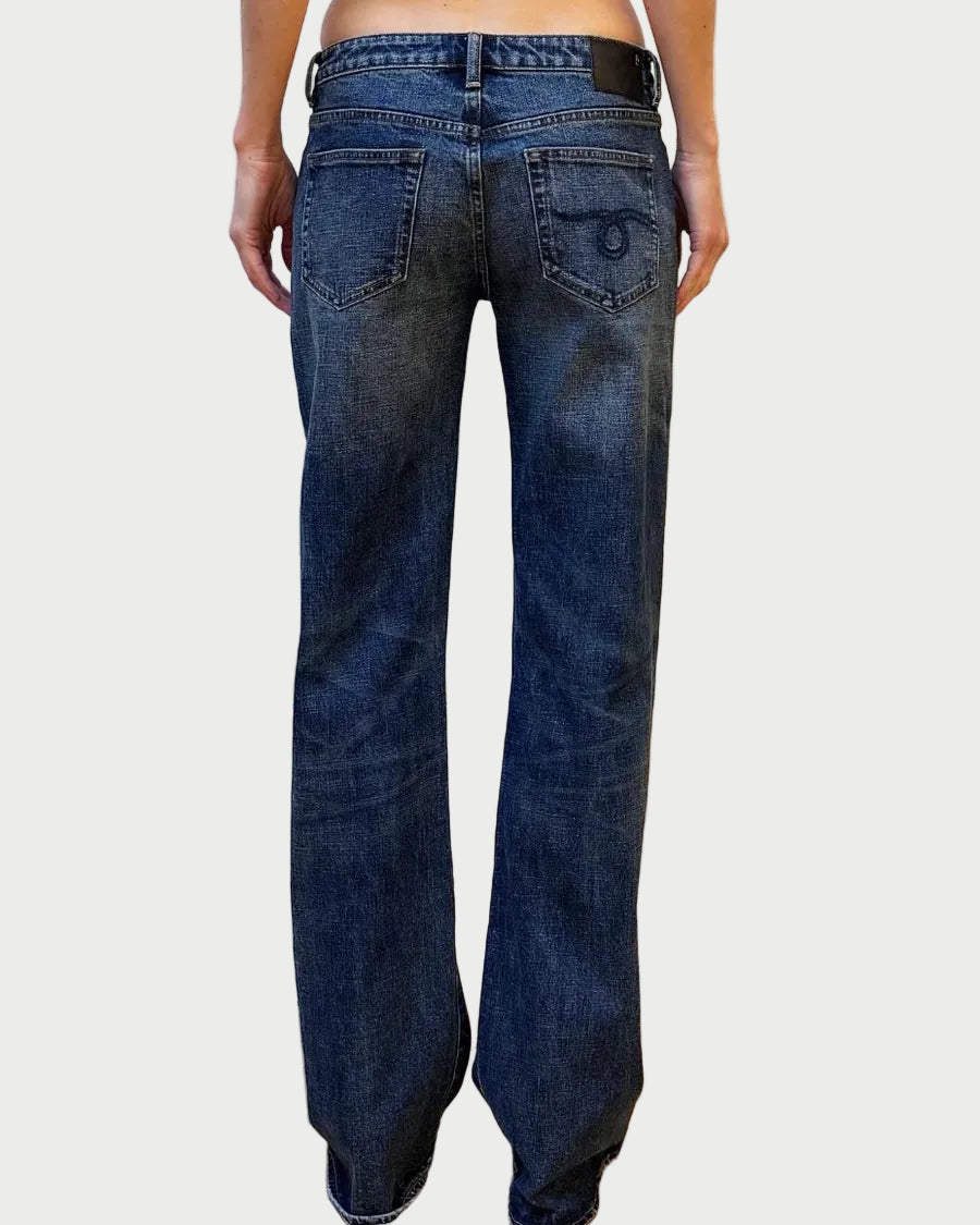 Rear view of a person wearing dark blue wide-leg denim jeans against a white brick wall and light wood floor.