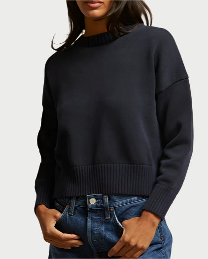 A woman wears the Janet Navy Cotton Sweater by perfectwhitetee—a loose, boxy, long-sleeve 100% cotton knit with ribbed cuffs and hem—paired with blue jeans. Her hands are in her pockets and her face is partially out of frame.