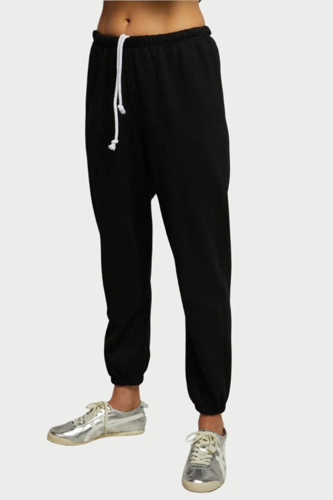 A person wearing the buttery soft Johnny Easy Sweatpants from perfectwhitetee, which feature an elastic waistband and white drawstring, stands against a neutral background. They are also sporting silver athletic shoes with white laces. The photo focuses on their lower body, not including the upper body or head.