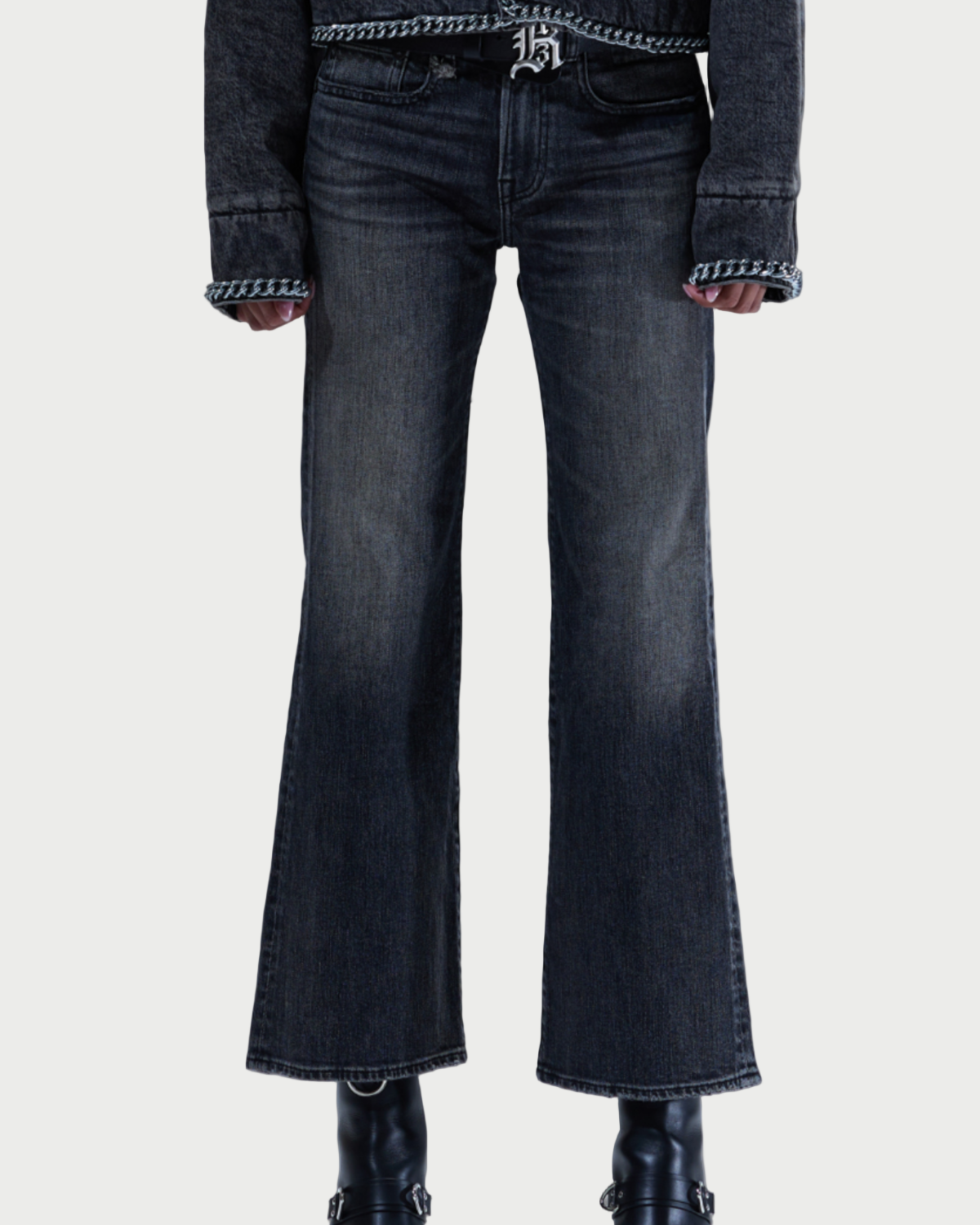 Someone wearing R13 Joan Kick Flare Jeans (dark wash) by R13, a dark long-sleeve top with patterned trim, and black boots stands against a plain light background.