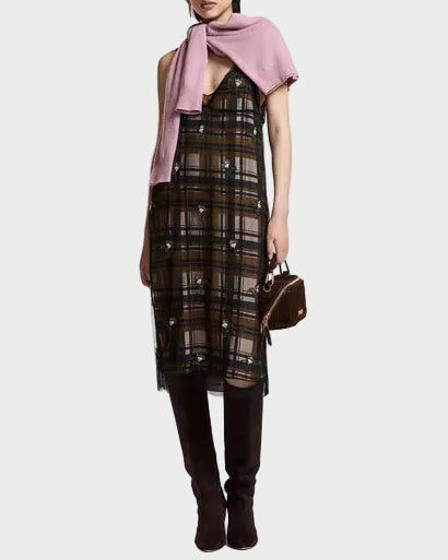 A model stands against a plain white studio background wearing a sheer black plaid midi dress with a pink shawl draped over her shoulders and dark brown knee-high boots, while holding a small brown handbag.