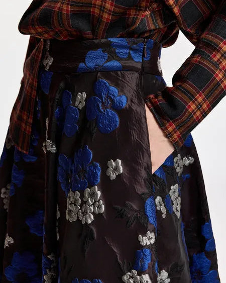 A person wearing a red plaid shirt and the Essentiel Antwerp Kathyanne Circle Midi Skirt—a shiny black skirt featuring blue and silver floral embroidery—with one hand in the skirt’s pocket.