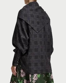 A person wearing the Essentiel Antwerp Kiss Oversized Shirt with Scarf—featuring a grey and black check pattern, large cuffs, a wide collar, and a detachable scarf—paired with a green skirt adorned with a pink floral pattern.