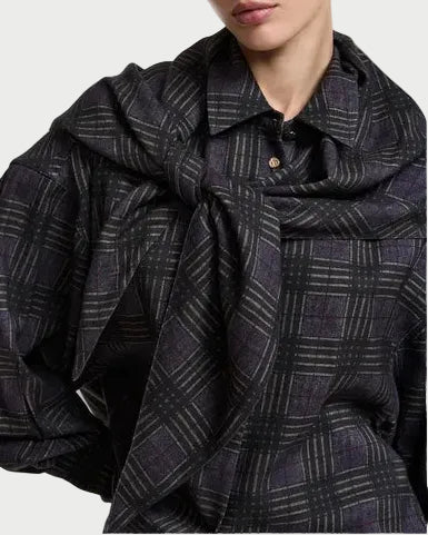 A person wears the Essentiel Antwerp Kiss Oversized Shirt w/Scarf by Essentiel Antwerp, featuring a dark plaid design with a subtle grey and black check pattern. The matching scarf is draped over their shoulders and tied in front, while a visible button near the collar adds detail to the understated look.