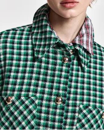 A close-up portrait of a model wearing a green and white checkered overshirt over a red and white plaid collared shirt, set against a plain light gray background.