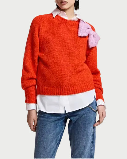 A model wearing a vibrant orange knit sweater with a pink bow detail on the shoulder, layered over a white collared shirt and paired with blue jeans, against a plain white studio background.