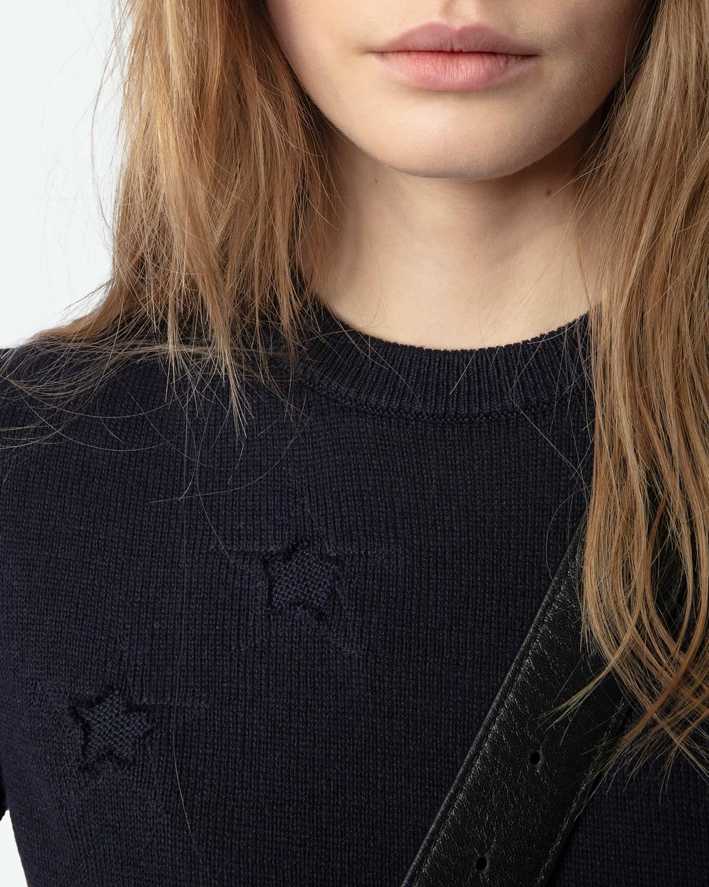 A partial view shows a person with long brown hair wearing the Zadig & Voltaire Short Sleeve Stars Sweater, characterized by distressed star intarsia cutouts. A black strap crosses their shoulder against a white backdrop, and their expression is neutral.