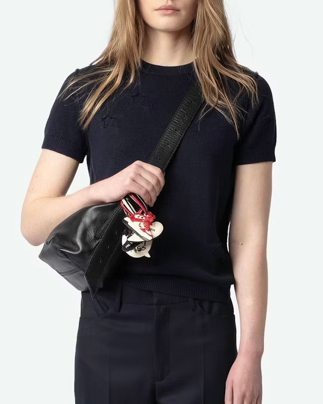 A woman in a black shirt and pants, accented by a colorful keychain on her handbag, stands against a neutral background. Her light brown hair cascades over her shoulders like the gentle weave of a Zadig & Voltaire Short Sleeve Stars Sweater.