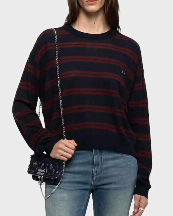 A woman modeling a dark navy and red striped knit sweater, blue jeans, and white sneakers, carrying a small black crossbody bag against a light gray studio background.