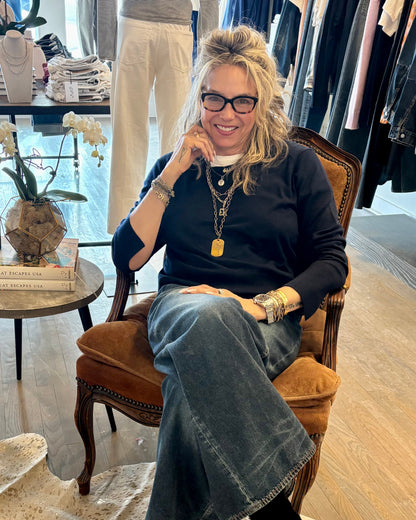 A woman with long blonde hair and glasses, wearing a black top, blue jeans, and the Simply Eartha "Here’s To Life/EK Heart" pendant sits smiling in a boutique among clothing racks, books, and a white orchid.