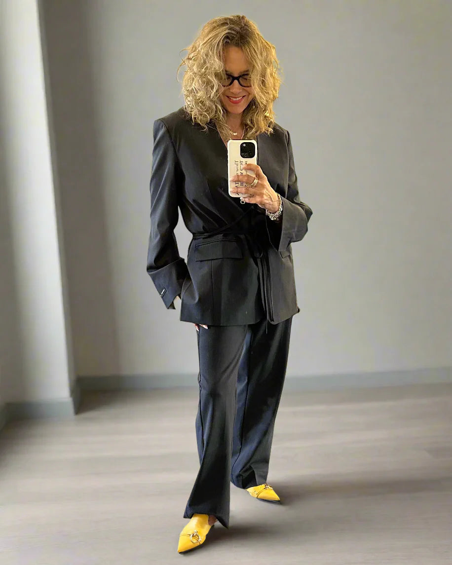 A person with curly blonde hair wears the oversized Herskind Indiana Blazer in black wool blend, smiling and taking a mirror selfie in a minimalist room with grey walls and floor, paired with yellow pointed shoes.