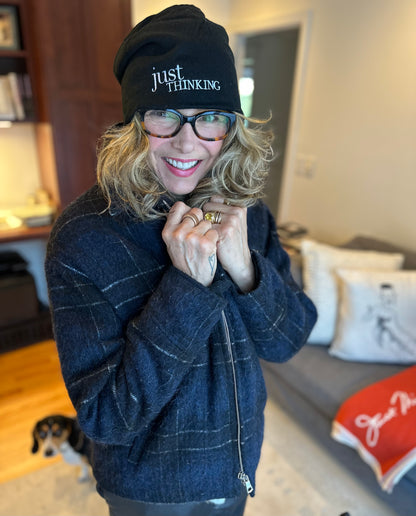 A woman with wavy blonde hair, glasses, and a Simply Eartha Slouchy "Just Thinking" Beanie smiles warmly, holding the collar of her dark, textured coat in the cozy winter warmth of a room with an inviting couch and blanket in the background.
