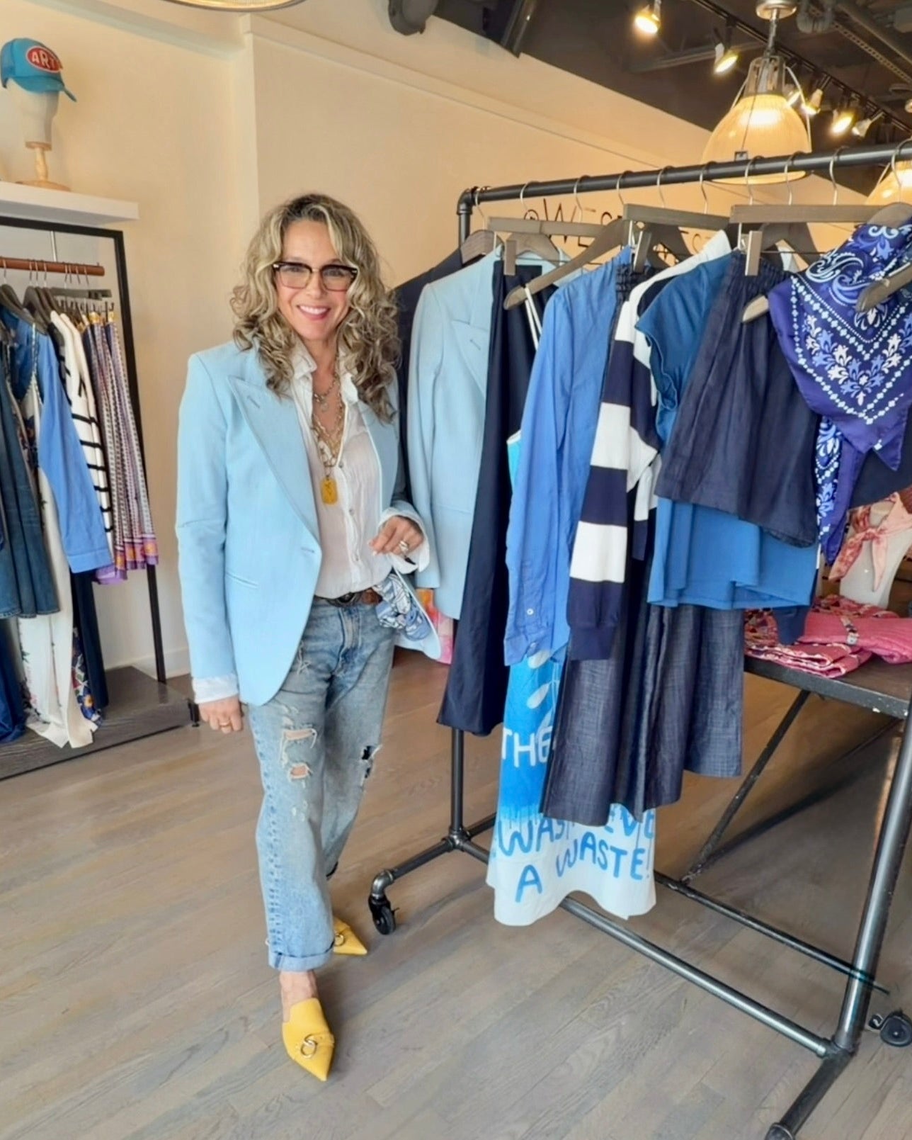 A smiling woman with wavy hair, sunglasses, a light blue blazer, and yellow shoes stands by a clothing rack wearing R13 Boyfriend Jean In Bain Blue by R13 in a stylish boutique.