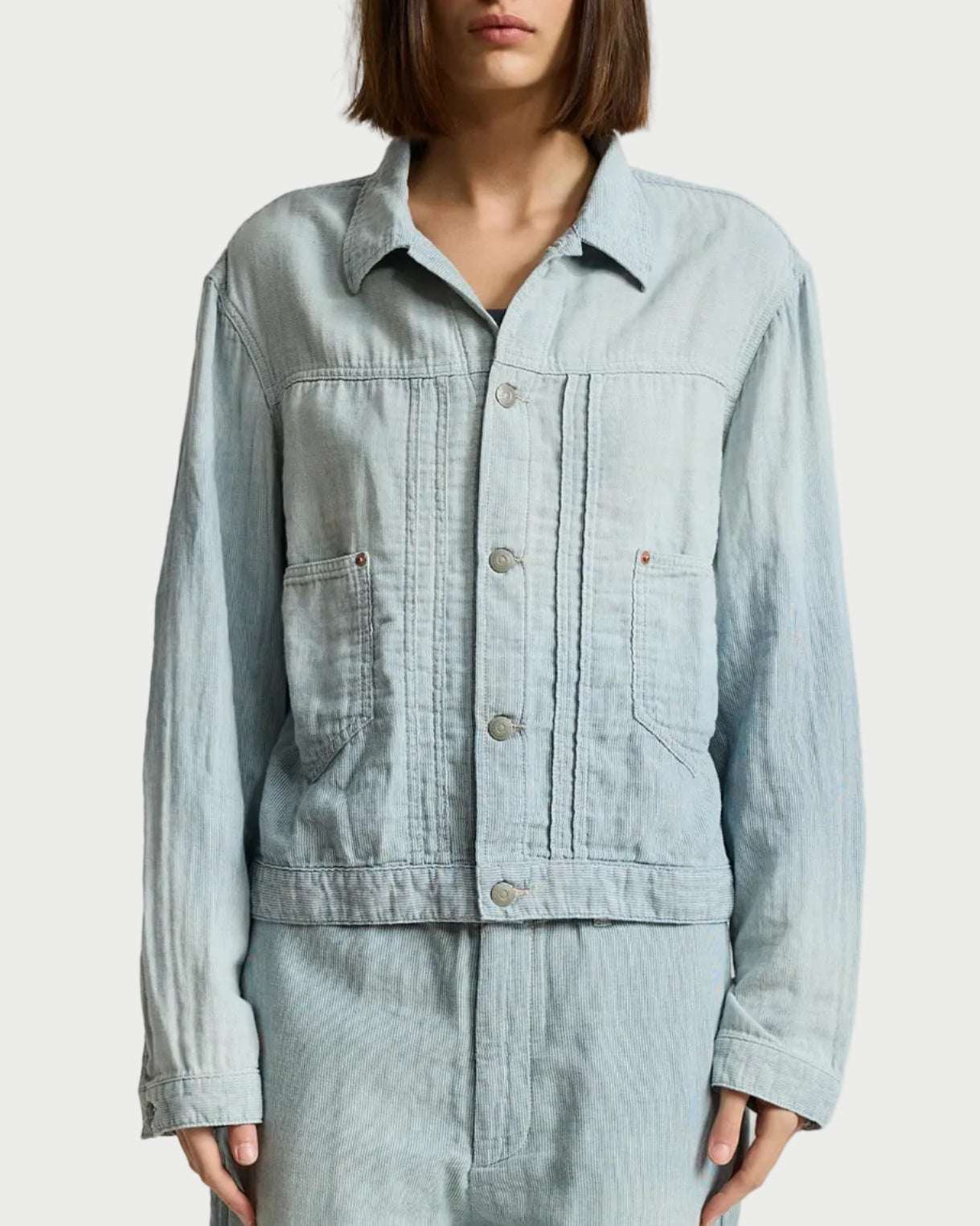 A model wearing a light blue denim utility jacket and matching light blue denim pants, standing against a neutral light gray studio background.