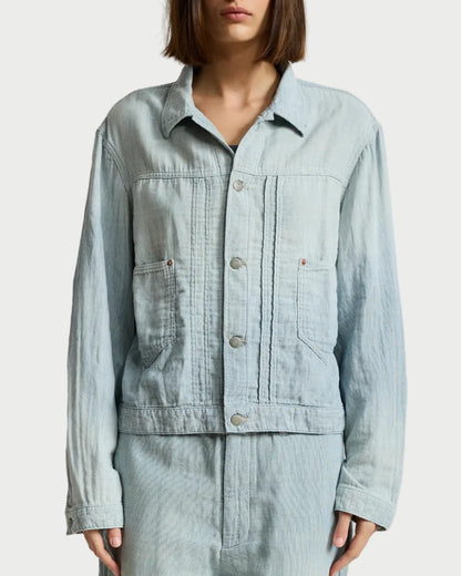 A model wearing a light blue denim utility jacket and matching light blue denim pants, standing against a neutral light gray studio background.