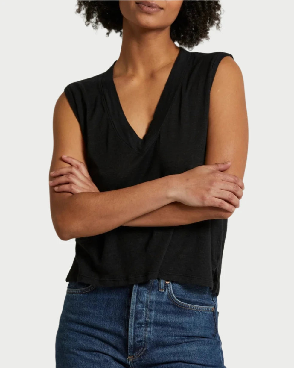 A person standing with arms crossed wears a Lucy V neck Tank in black from perfectwhitetee and blue jeans. The background is plain and light-colored. The person's face is not fully visible.