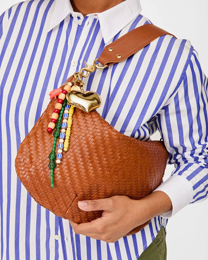 A person in a blue and white striped shirt holds CLARE V.'s Le Long Beaded Tassel, a brown woven shoulder bag adorned with colorful beads, a gold heart-shaped charm, and a playful beaded tassel.