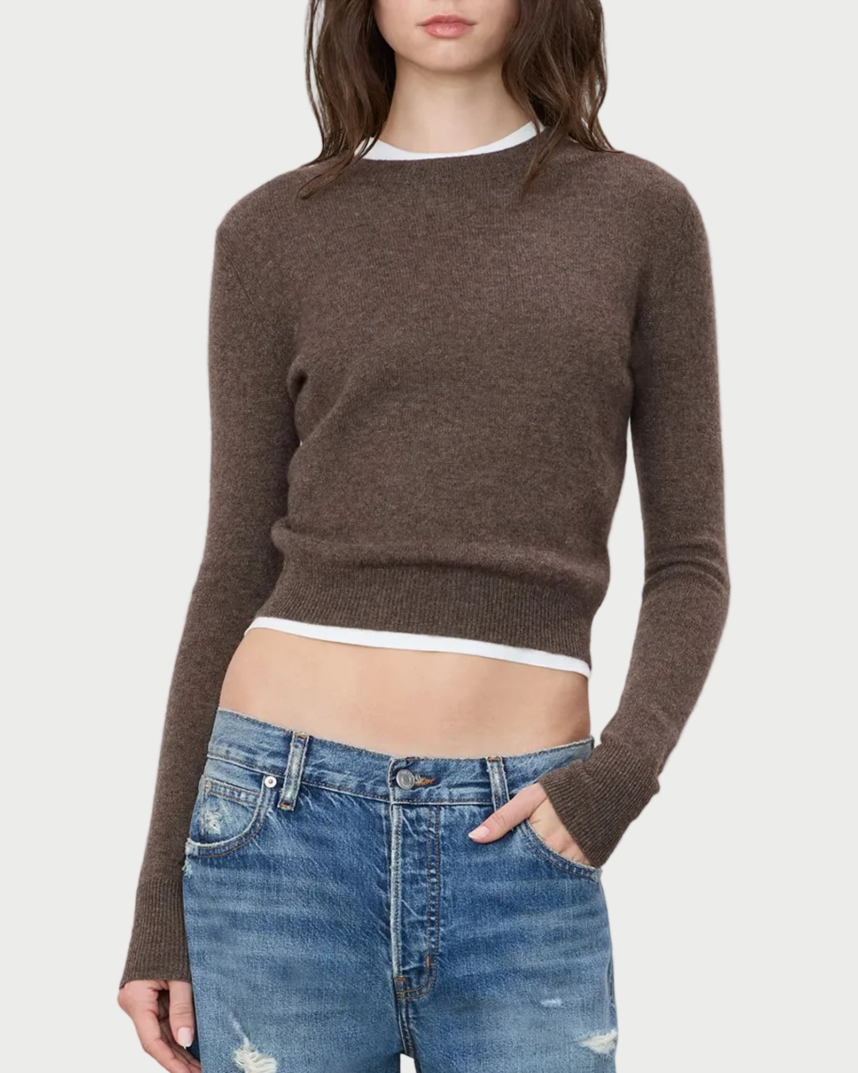 A woman wearing the ÉTERNE Francis Sweater by Eterne in brown, layered over a white shirt and paired with blue distressed jeans, stands with her right hand in her pocket against a plain background.