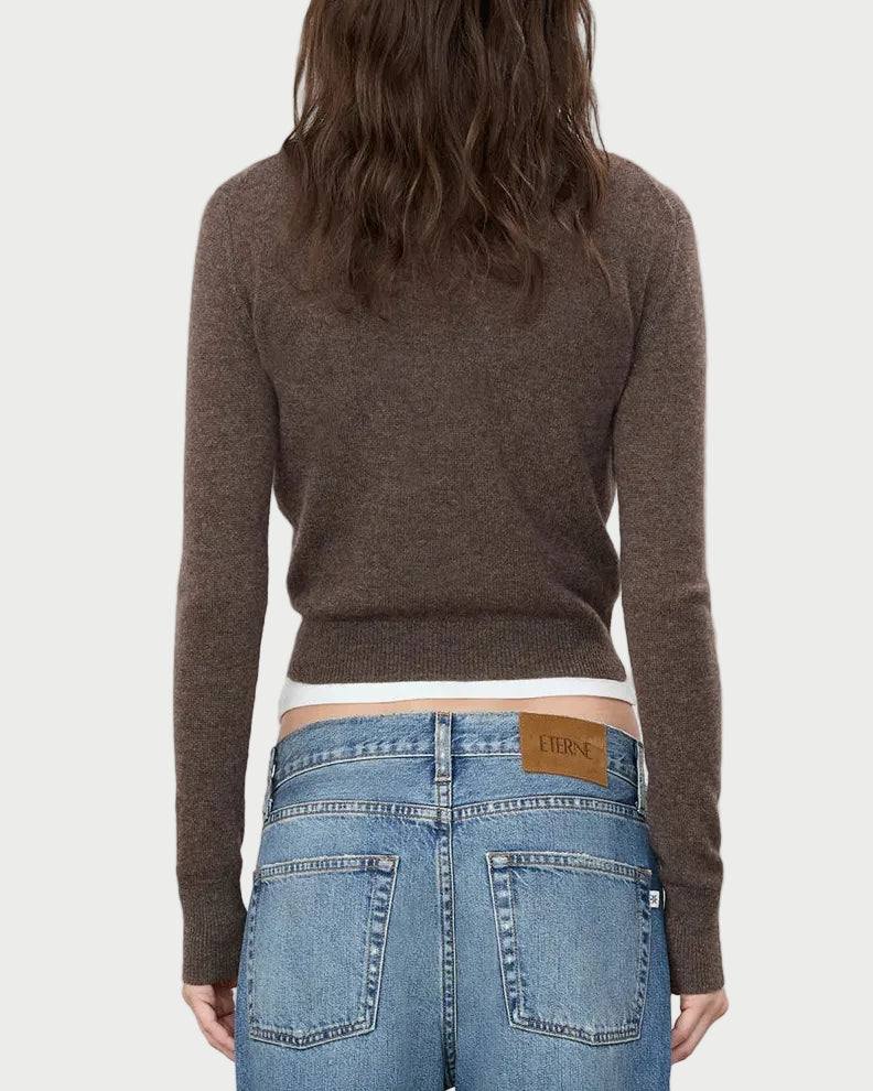 A person with wavy brown hair, seen from the back against a plain light gray background, wears the Eterne ÉTERNE Francis Sweater—a cropped cashmere piece—paired with blue jeans featuring a tan leather “ÉTERNE” patch.