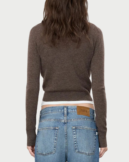 A person with wavy brown hair, seen from the back against a plain light gray background, wears the Eterne ÉTERNE Francis Sweater—a cropped cashmere piece—paired with blue jeans featuring a tan leather “ÉTERNE” patch.