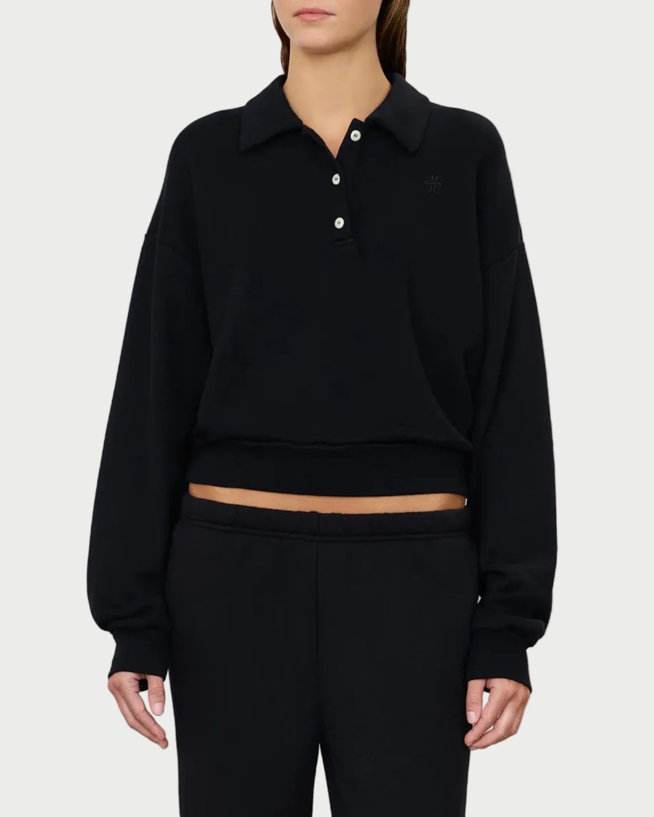 A person wearing the Éterne Cropped Polo Sweatshirt by Eterne—a relaxed fit, black polo sweatshirt featuring three buttons and a slightly cropped length that reveals a hint of midriff—is paired with matching black pants and stands against a plain light background.