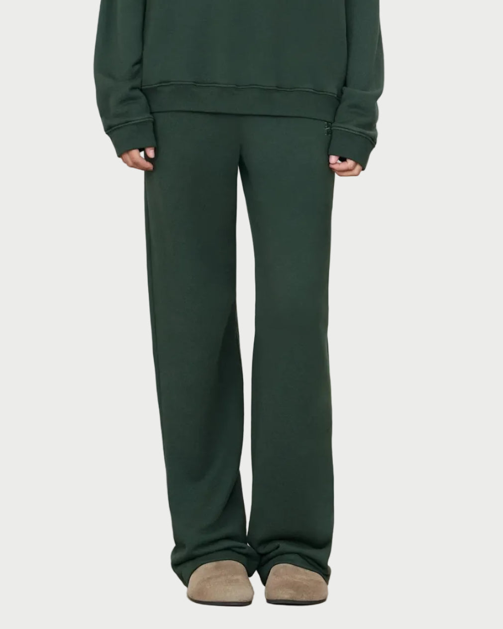 A person wearing Éterne Oversized Wide Leg Sweatpants by Eterne in dark green, paired with a matching sweatshirt and beige slippers, stands against a plain background with only the lower half of the body visible.