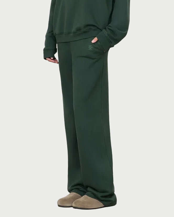 Wearing the Éterne Oversized Wide Leg Sweatpant by Eterne in dark green, paired with a matching sweatshirt and brown slippers, the person stands with one hand in their pocket against a plain background.