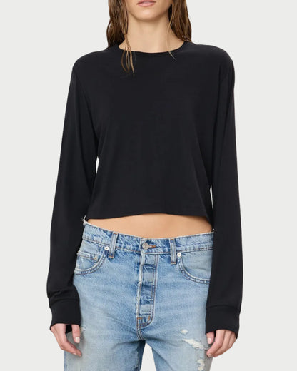 A person is standing against a plain background wearing the Éterne Long Sleeve Boxy Tee by Eterne in a soft cotton-modal blend, paired with light blue ripped jeans. Their look is highlighted by a black cropped silhouette top, and their hair appears wet with their face partially out of frame.