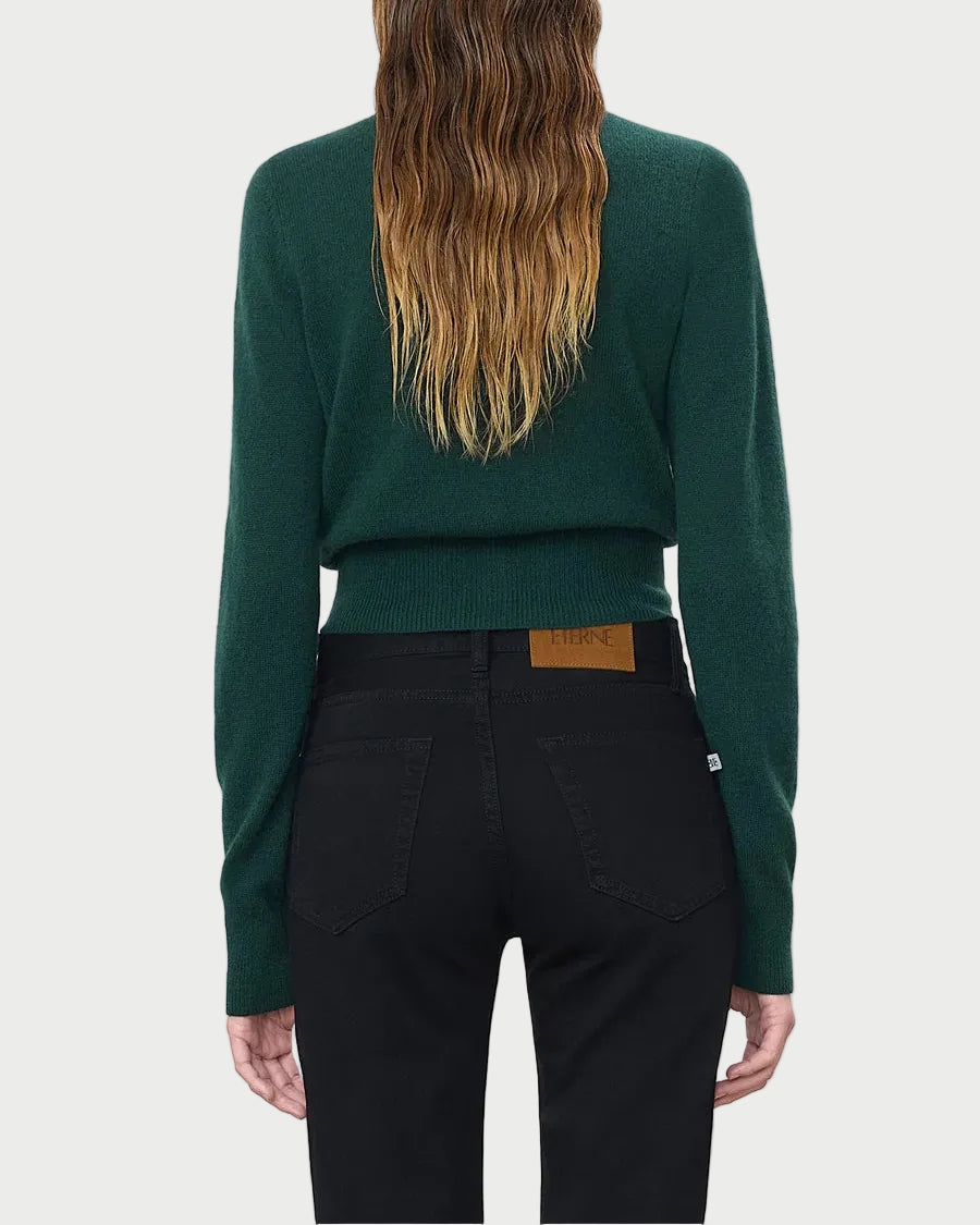 A person with long, wavy brown hair is shown from behind against a plain light background, wearing the Eterne Éterne Reese Cashmere Sweater in dark green slim fit paired with black jeans.
