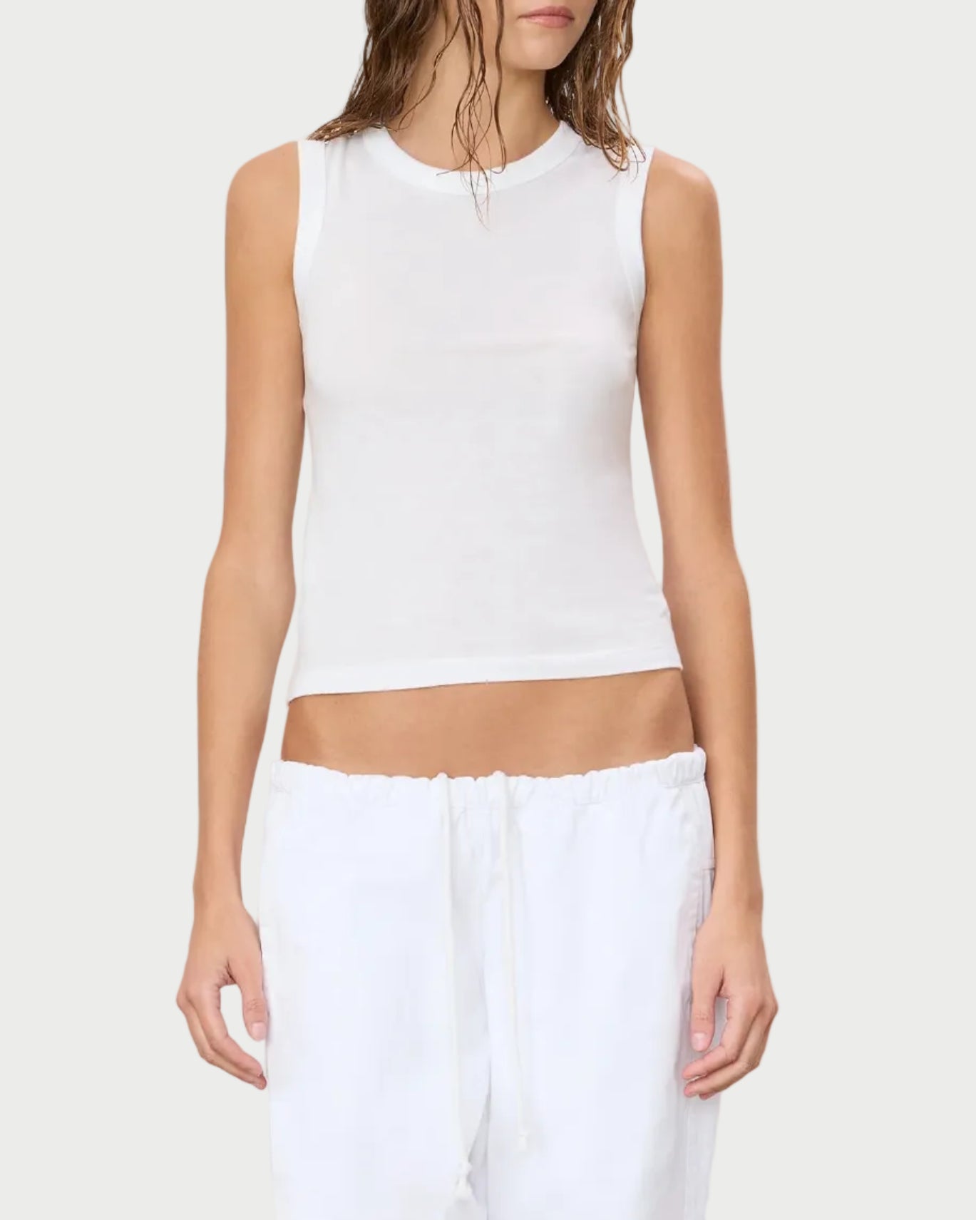 A person stands against a plain light background, wearing the Eterne Éterne Fitted Muscle Tank—a slim fit, sleeveless white cotton-modal blend top—paired with white drawstring pants. Their head is partially out of frame, and they have wet, wavy hair.