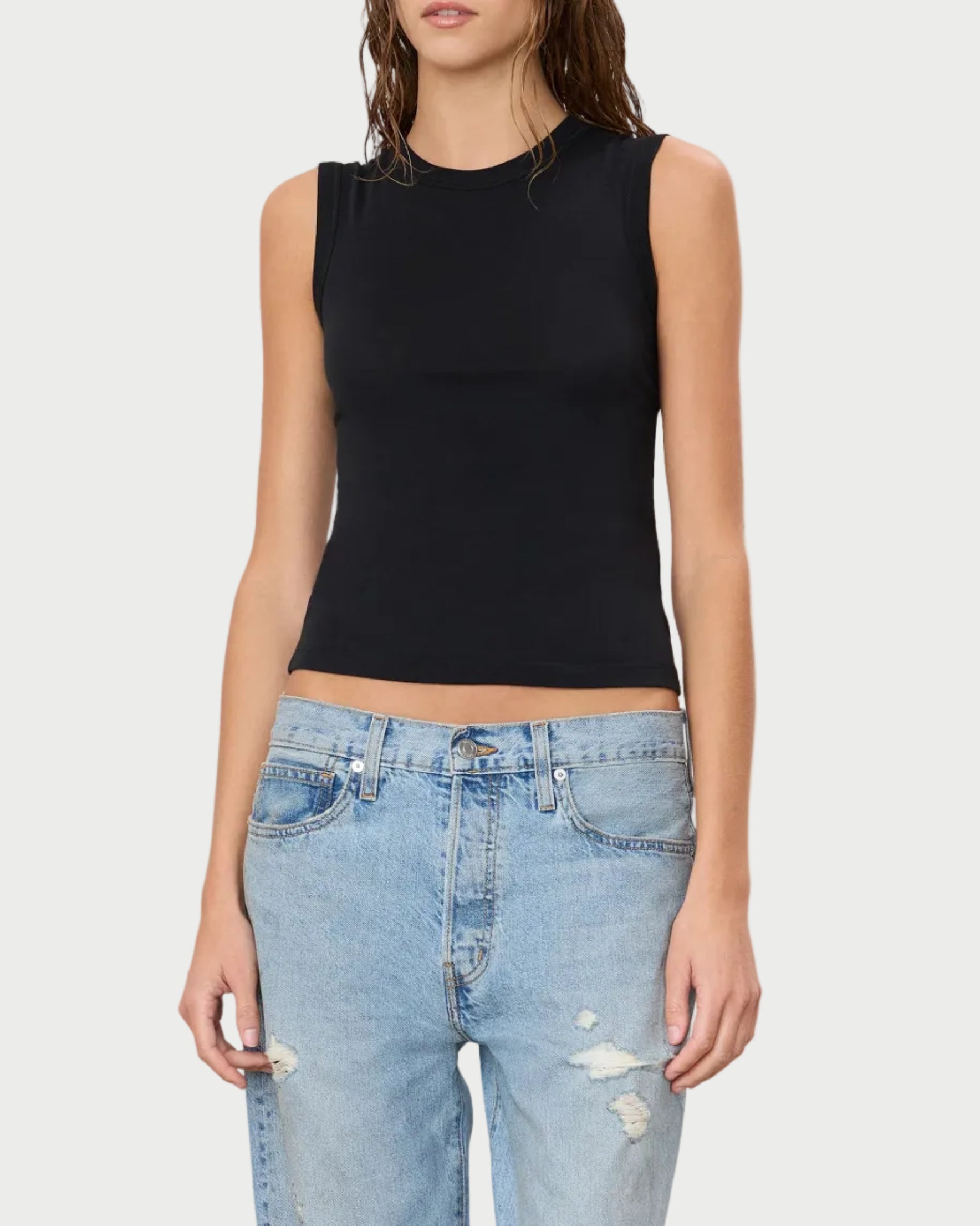 A woman wearing the Éterne Fitted Muscle Tank by Eterne—a sleeveless black top crafted from a soft cotton-modal blend—and light blue ripped jeans stands against a plain white background.