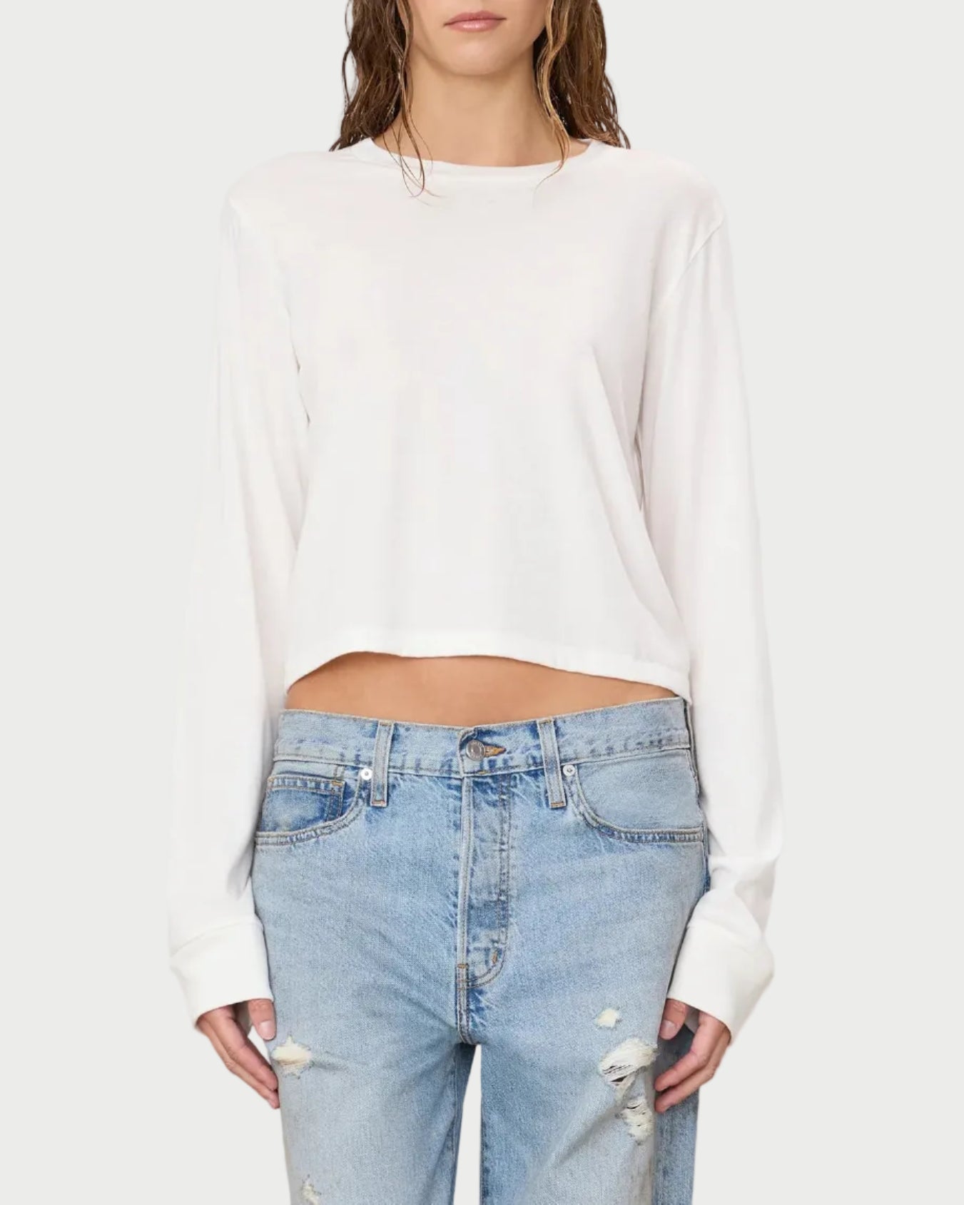 A woman wears the Éterne Long Sleeve Boxy Tee by Eterne, featuring a relaxed boxy fit in a soft cotton-modal blend, paired with light blue ripped jeans and standing against a plain background.