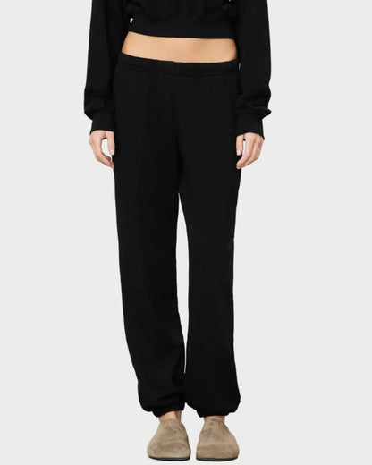 Wearing the Eterne Boyfriend Pocket Sweatpant in black French terry, paired with a black long-sleeve top and beige slippers, the individual stands against a plain light background. Only the body from shoulders to feet is visible; the face is not shown—showcasing an effortlessly elevated loungewear look by Eterne.