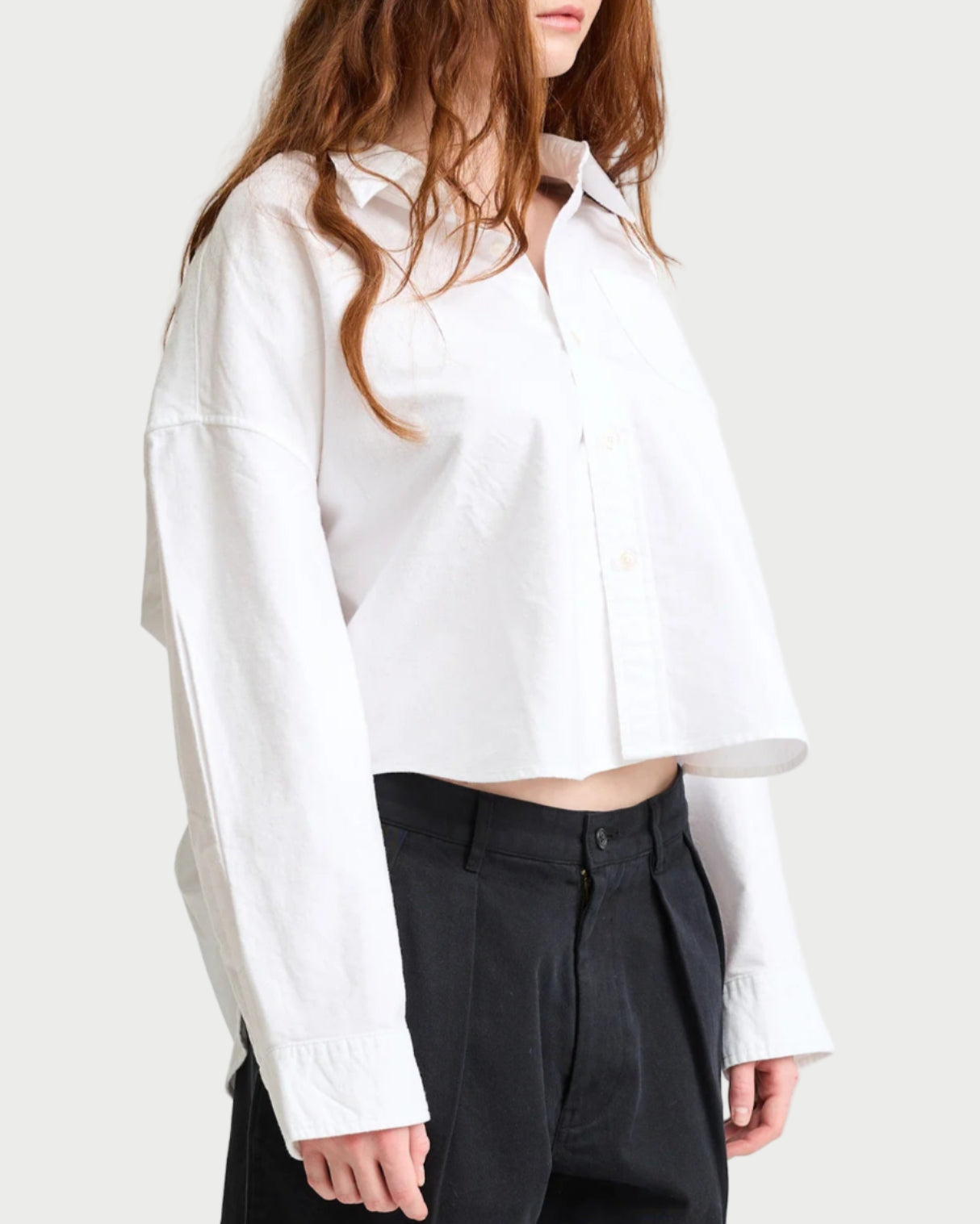 A person with long, wavy brown hair wears the Denimist Cropped Button Front Shirt by Denimist and black pants against a plain light background. The loose-fitting white shirt offers a modern look with its slightly revealed midriff.