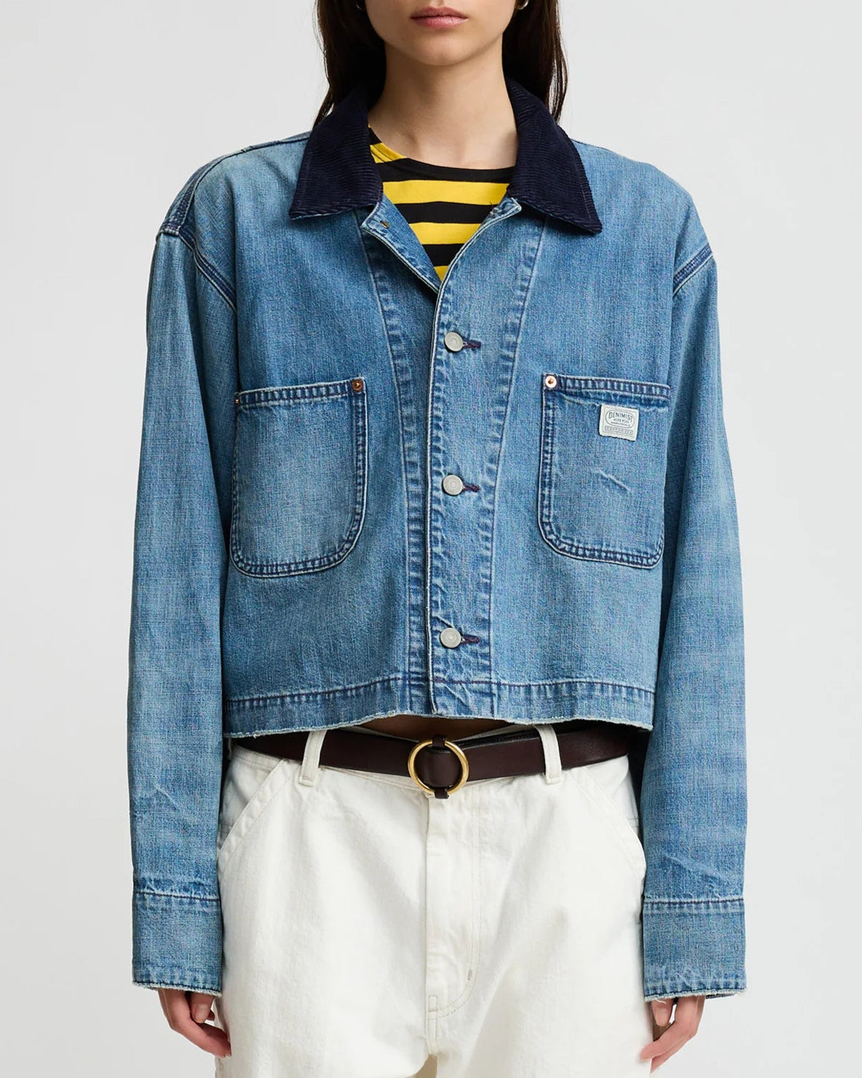 A person wears the Denimist Chore Denim Jacket—a cropped, structured cotton piece with large front pockets and a black collar—layered over a yellow and black striped shirt, white pants, and a brown belt with a gold buckle.