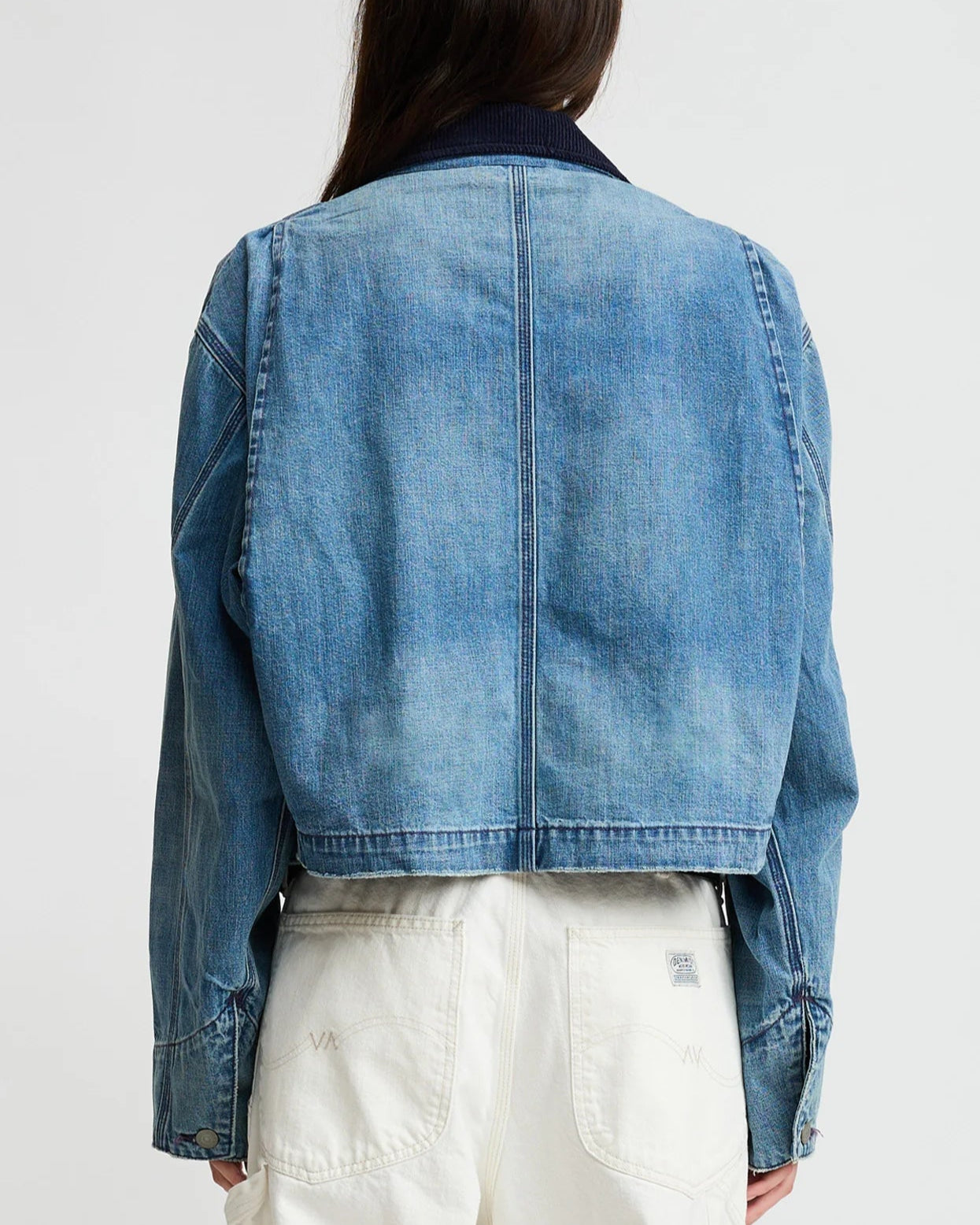 A person with long hair is seen from behind wearing the Denimist Chore Denim Jacket and high-waisted white pants against a plain background.
