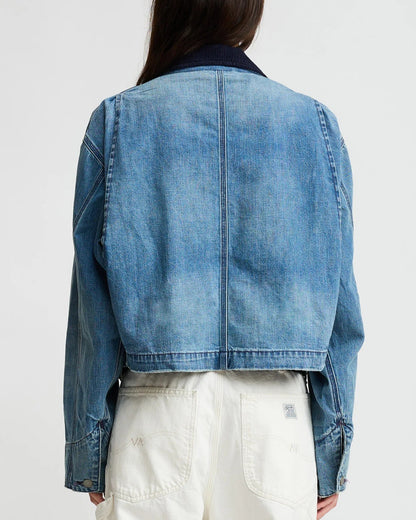 A person with long hair is seen from behind wearing the Denimist Chore Denim Jacket and high-waisted white pants against a plain background.