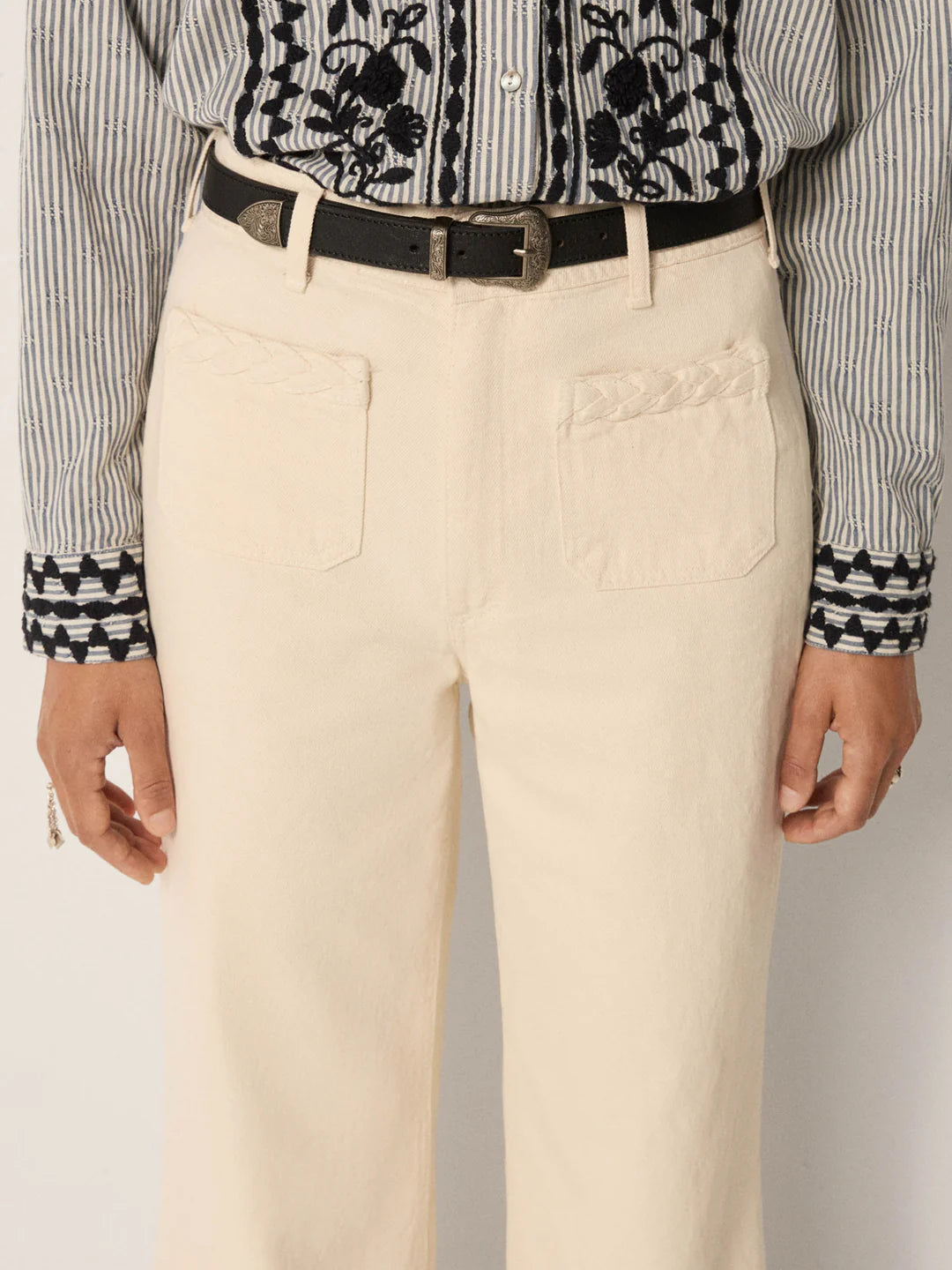 A person wears Maison Hotel Hamlet Pants—high-waisted, cream-colored with front pockets—paired with a black belt and a black-and-white striped shirt featuring floral and geometric embroidery on the cuffs and chest. Only torso and upper legs are visible.