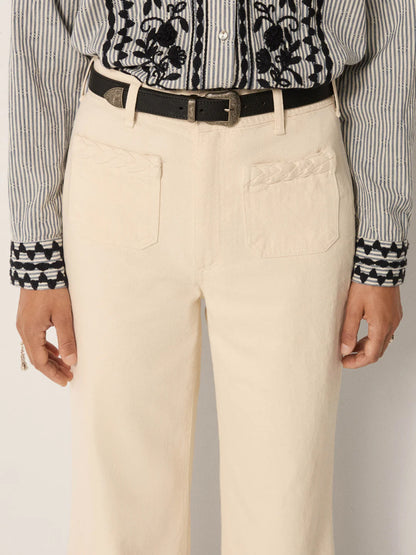 A person wears Maison Hotel Hamlet Pants—high-waisted, cream-colored with front pockets—paired with a black belt and a black-and-white striped shirt featuring floral and geometric embroidery on the cuffs and chest. Only torso and upper legs are visible.