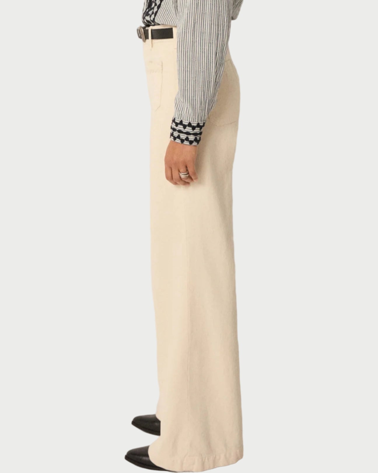 A person stands sideways in a striped shirt tucked into Maison Hotel Hamlet Pants—high-waisted, wide-leg cream trousers made from recycled cotton—styled with black pointed-toe shoes against a light background.